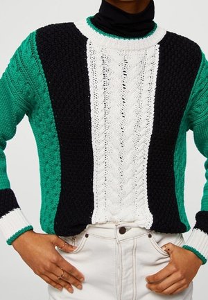 Personne portant un pull torsadé vert, noir et blanc avec un pantalon blanc, les mains posées sur les hanches.