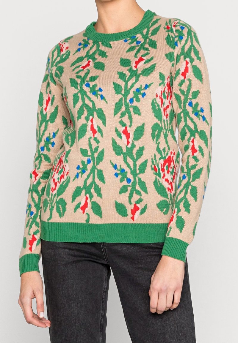 Pull en tricot beige avec des motifs floraux vibrants en vert, rouge et bleu. Il possède un col rond et des poignets et un ourlet côtelés en vert.