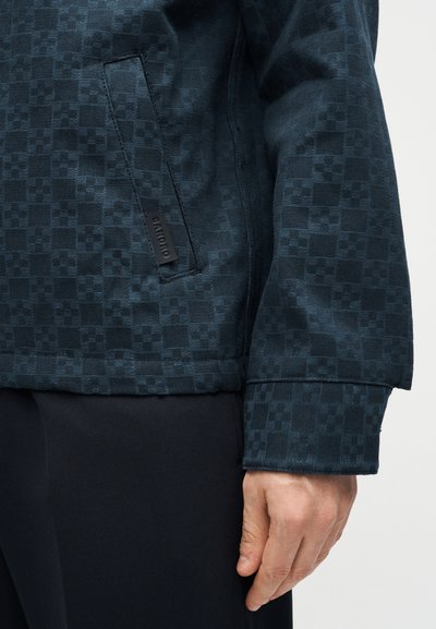 Σκούρο σακάκι με μοτίβο και μπροστινή τσέπη που δείχνει μια μικρή μαύρη ετικέτα, φορεμένο με μαύρο παντελόνι και εμφανές δεξί χέρι πάνω σε απλό φόντο.