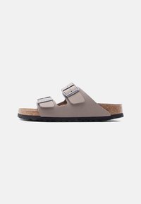 Μπεζ σανδάλι Birkenstock με δύο λουριά και ασημένια αγκράφες, πάτο από φελλό και μαύρη υφή στην σόλα, εμφανίζεται σε πλευρική όψη σε λευκό φόντο.