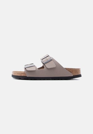 Sandalia beige Birkenstock de dos tiras con hebillas plateadas, plantilla de corcho y suela negra texturizada, vista de perfil sobre fondo blanco.