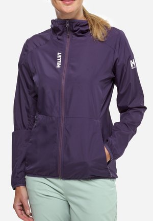 Femme portant une veste Millet violette zippée avec des logos sur la poitrine et la manche, pantalon clair, debout les mains dans les poches.