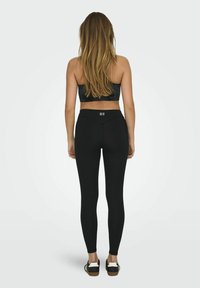 Zwarte legging van rekbaar materiaal, met een hoge tailleband en een naadloos ontwerp. G gedragen met een zwarte sportbeha en casual schoenen.