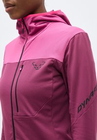 Κοντινό πλάνο ενός ατόμου που φοράει διχρωμία μωβ ζακέτα με φερμουάρ και κουκούλα, με τσέπη στο στήθος και το λογότυπο "DYNAFIT" στο μανίκι.