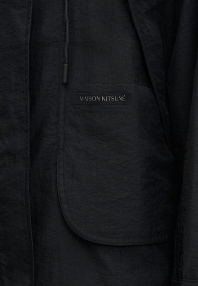 Veste noire texturée avec une poche avant portant une étiquette "MAISON KITSUNÉ" et un cordon noir visible.