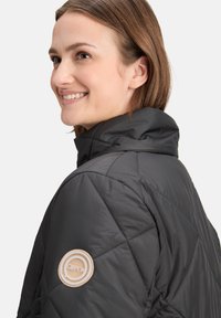 Sort quiltet jakke med høj krave, der har et rundt logo-patch på venstre ærme og en glat, blank overflade.