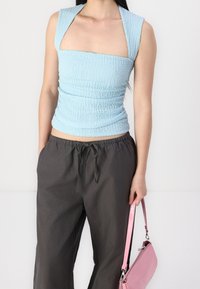 Vrouw draagt lichtblauw, gestructureerd topje en donkergrijze broek, houdt een kleine roze handtas met polsbandje vast.