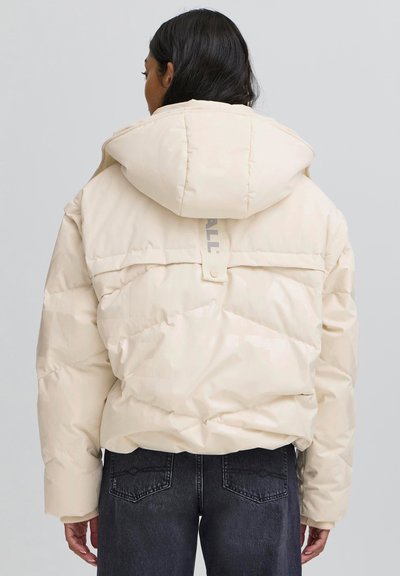 Bēšs pūkains jaciņš ar kapuci, dekorēts ar kapitonēta rakstu, elastīgām aprocēm un aizmugurējo zīmola uzlīmi; valkāts kopā ar tumšām džinsām.