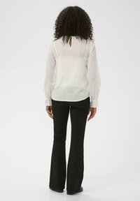 Blouse blanche avec des manches longues et bouffantes, en tissu texturé, et une ouverture en forme de clé à l'arrière. Associée à un pantalon noir évasé.