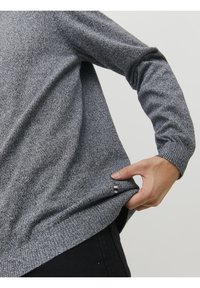 Pull gris texturé à manches longues, avec poignets et ourlet côtelés. Une petite étiquette d'accent est visible près du bord inférieur. Matière tricotée douce.