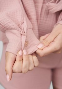 Tessuto rosa chiaro, con una texture e un dettaglio a bottone sul polsino. Il design presenta una chiusura a cravatta ed è progettato per raccogliersi leggermente sul polso.
