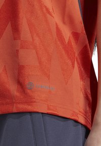 T-shirt de sport à motif rouge avec une finition texturée, arborant le logo "AEROREADY" bleu près de l'ourlet. Porté avec un short marine.