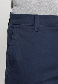 Tmavě modré bavlněné kalhoty s rovně střiženým designem, plochým předkem, poutky na pásek a bočními kapsami. Hladká textura s jemným prošíváním.