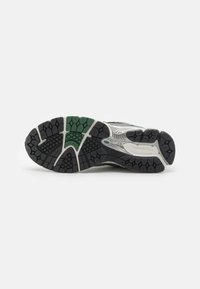 Melna un zaļa skriešanas apavu zolīte ar teksturētu gumiju, kurai ir seškantīga raksta izskats un sekli urbumi, lai nodrošinātu saķeri, ar marķējumu "newbalance".