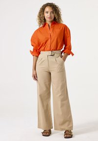 Oranje overhemd met opgerolde mouwen, gecombineerd met beige hoge wijde broek en bruine sandalen met decoratieve accenten.