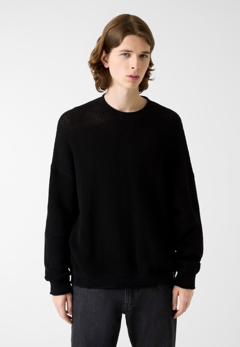 Schwarzer Strickpullover mit strukturiertem Muster, rundem Ausschnitt und lockerem Schnitt. Lange Ärmel mit gerippten Bündchen. Kombiniert mit dunklen Jeans.