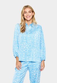 Kék mintás ing hosszú puffos ujjakkal és gallérral, bonyolult fehér minták díszítik. Hozzá illő kék mintás nadrággal párosítva.