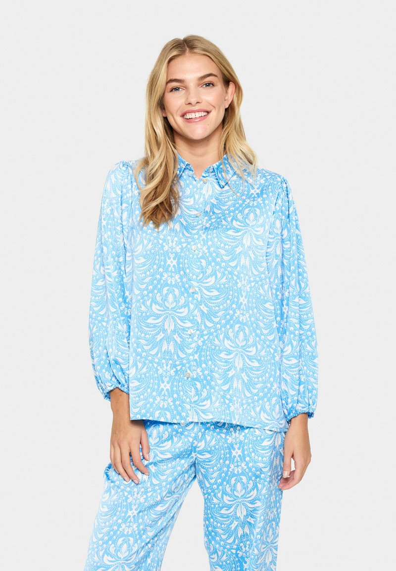 Kék mintás ing hosszú puffos ujjakkal és gallérral, bonyolult fehér minták díszítik. Hozzá illő kék mintás nadrággal párosítva.