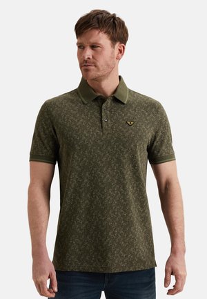 Hombre con cabello corto castaño que lleva una camisa tipo polo de color verde oliva con patrón, cuello y mangas cortas, posando frente a un fondo blanco.