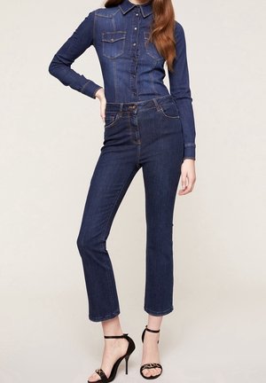 Kobieta ubrana w ciemnoniebieską dżinsową koszulę na guziki i dżinsy, w połączeniu z czarnymi sandałami na wysokim obcasie, pozująca z ręką na biodrze.