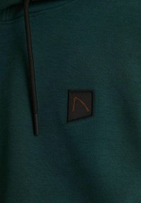 Felpa verde realizzata in morbido tessuto, con una toppa rettangolare nera con un design geometrico arancione e cordini neri.