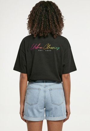 Person med krøllet hår iført sort oversized t-shirt med teksten "Urban Classics EST. 2005" i flere farver og lyseblå opkrogede denimshorts.