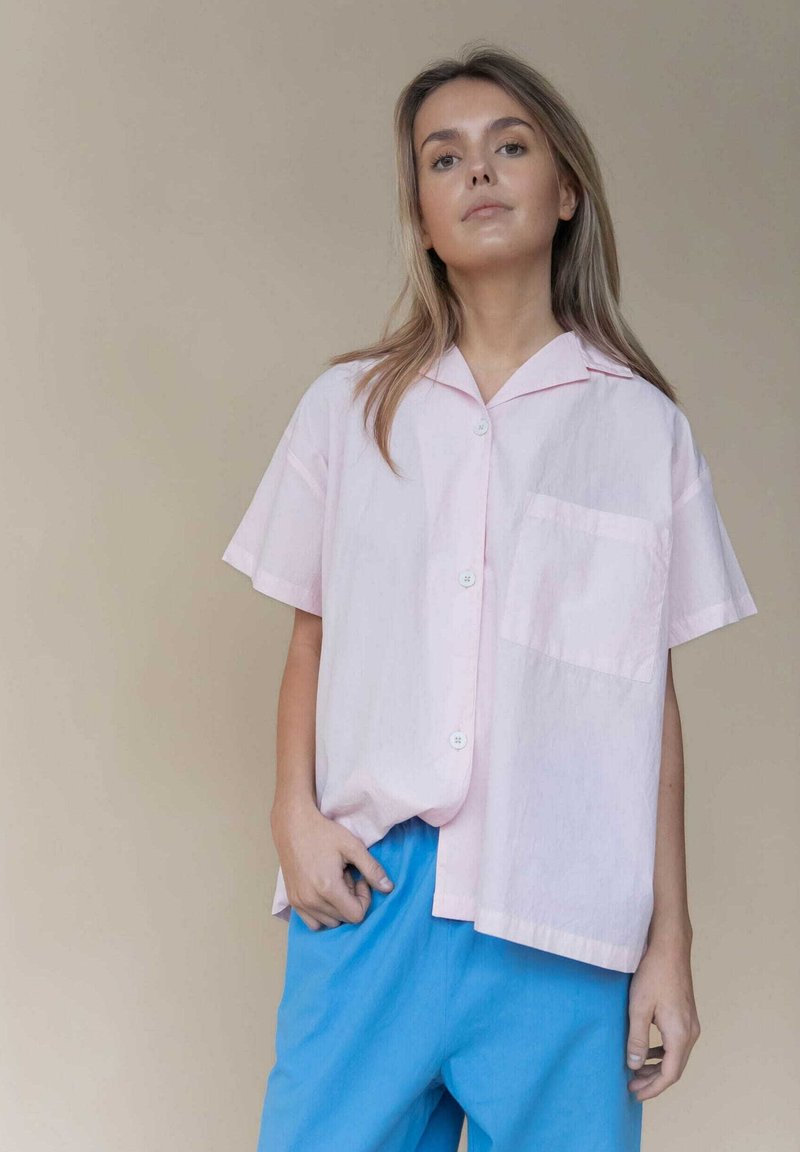 Korte mouwen, lichtroze overhemden met knopen en een zak, gecombineerd met blauwe loszittende broeken, tegen een neutrale achtergrond.