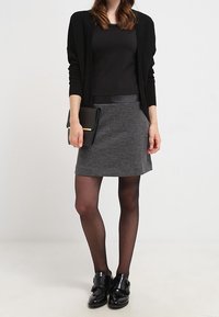 Černý cardigan s dlouhým rukávem, černý top, šedá sukně s jemnou texturou, černé punčocháče a černé boty na přezku. Drží černou kabelku.