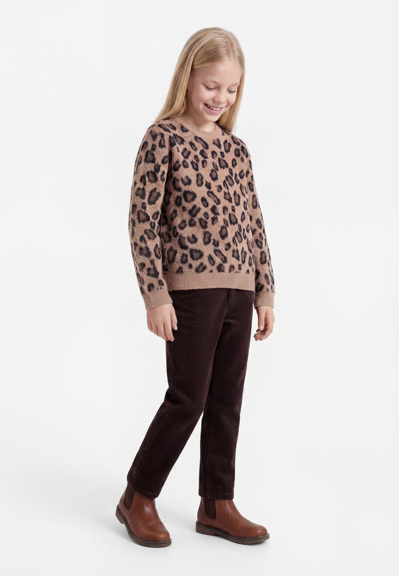 Fille portant un pull à motif léopard, un pantalon en velours côtelé marron foncé et des bottines marron, debout et regardant vers le bas avec un sourire.