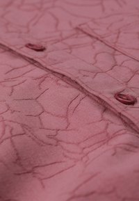 Dengtos rožinės spalvos audinys su subtiliu įtrūkimų raštu, turintis mygtukų užsegimus ir lygų paviršių. Medžiaga atrodo minkšta ir lengva.