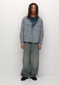 Fiatal személy hosszú rasztás hajjal, kockás ingben, grafikás pólóban, túlságosan nagy, kifakult farmernadrágban és fekete cipőben áll egy sima háttér előtt.