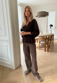 Braunes Wickeloberteil mit weiten Ärmeln, kombiniert mit braunen, locker sitzenden Hosen und hellgrauen Slippern. Hält eine weiße Tasse.