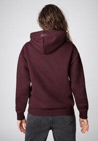 Bordeauxhoody met een grote capuchon, geribbelde manchetten en tailleband. Zacht materiaal; heeft een logolabel aan de achterkant van de nek. Gedragen met donkergrijze jeans.