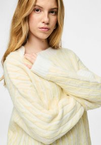 Storas, kreminės spalvos megztinis su vertikaliomis geltonomis ir baltomis juostelėmis, plačia iškirpte ir ilgomis rankovėmis.