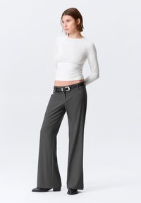 Haut blanc ajusté à manches longues, associé à un pantalon gris à jambes larges, présentant une texture lisse et une ceinture noire avec une boucle argentée.