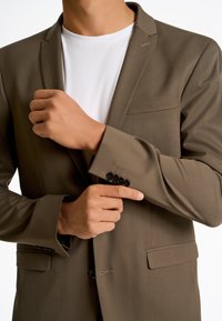 Blazer marrone con revers, taschino sul petto singolo e polsini con bottone. Sotto, una camicia bianca a girocollo è visibile. Tessuto dalla superficie liscia.