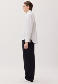 Witte langemouwen overhemd van structuurstof, gecombineerd met donkere, wijde broek en witte sneakers, die een minimalistisch ontwerp tonen.