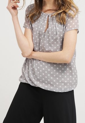 Helle graue Bluse mit Punkten aus durchsichtigem Stoff, mit kurzen Ärmeln und einem Schlüsselloch-Ausschnitt. Kombiniert mit schwarzen, locker sitzenden Hosen.