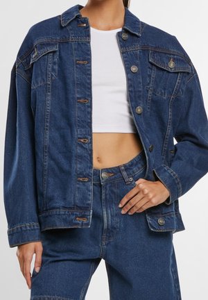 Frau trägt dunkelblaue Jeansjacke und passende Jeans mit weißem bauchfreiem Tanktop, Hand ruht in der Hosentasche.