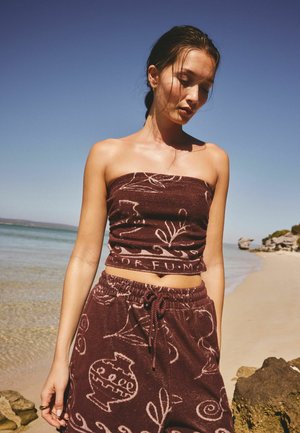 Mladá žena stojí na piesočnatej pláži, oblečená v hnedom strapless topu a zodpovedajúcich šortkách s bielymi abstraktnými vzormi.