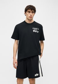 Mladi moški, oblečen v črno majico s kratkimi rokavi z belim besedilom in ikonami zemeljskega kroga ter črne kratke hlače Nike z belimi stranskimi črtami, stoji pred enobarvnim ozadjem.