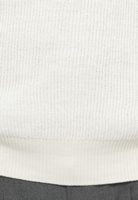 Πλεχτό πουλόβερ σε λευκό χρώμα με υφή, διαθέτοντας ριχτό τελείωμα. Συνδυάζεται με γκρι ύφασμα που δείχνει λείο φινίρισμα.