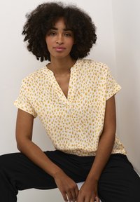 Kortærmet bluse i gul med multikolor abstrakt mønster, med v-hals design og en løstsiddende pasform, parret med sorte bukser.