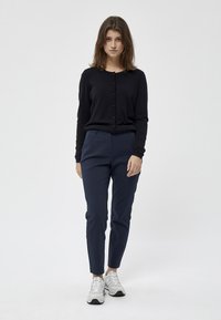 Cardigan lungo nero con maniche a sbuffo e bottoni, abbinato a pantaloni navy slim-fit. Il modello indossa scarpe da ginnastica grigie. Design semplice e sartoriale.