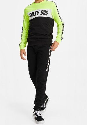 Chlapec v černé a neonově zelené mikině a černých kalhotách s nápisem "Salty Dog", kráčející v černobílých teniskách.