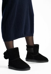 Bottines noires en daim avec une texture douce, dotées d'un accent pompon et de détails à rayures horizontales à l'arrière. Semelle plate.