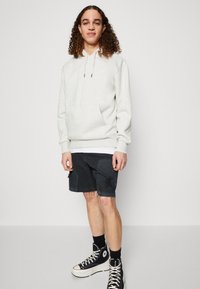 Felpa grigia chiara con stringhe e tasca frontale, abbinata a pantaloni corti neri strappati e sneakers alte nere. Look casual, stile rilassato.