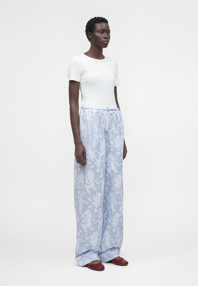 Γυναίκα όρθια που φοράει λευκό κοντομάνικο τοπ, άνετο ανοιχτό μπλε παντελόνι με λευκά λουλουδάτα σχέδια και κόκκινα επίπεδα παπούτσια μπροστά σε απλό φόντο.