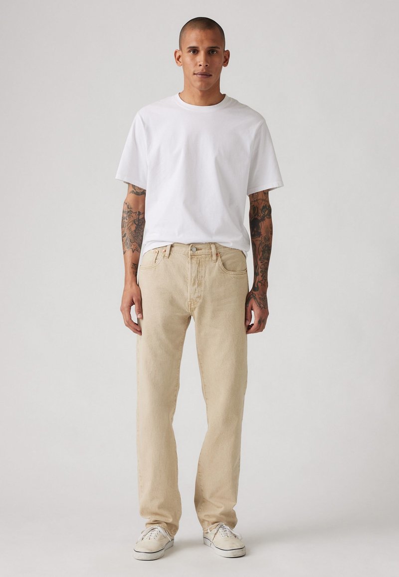 Beige Jeans im geraden Schnitt, kombiniert mit einem schlichten weißen T-Shirt. Merkmale sind ein Frontknopfverschluss und Taschen. Weiße Sneakers mit Gummisohle.