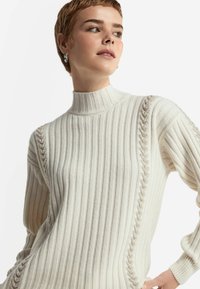 Cremefarvet ribstrikket turtleneck sweater med en struktureret kabeldetalje langs skuldersømmene. Tætsiddende stil med lange ærmer.